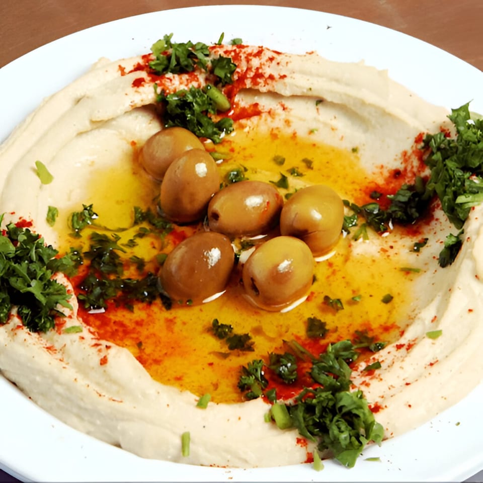 Creamy hummus topped with green olives, olive oil, paprika, and fresh parsley on a white plate.